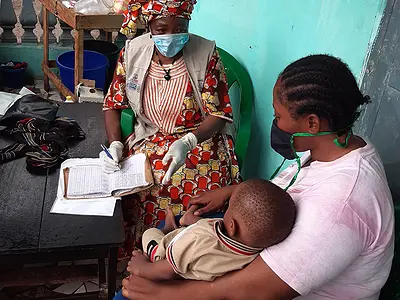An African health worker wearing a surgical mask talks with a mother and a young boy.