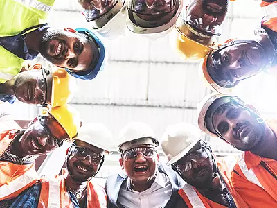 A group of engineers in hard hats at an Indian plant form a circle and smile for the camera.