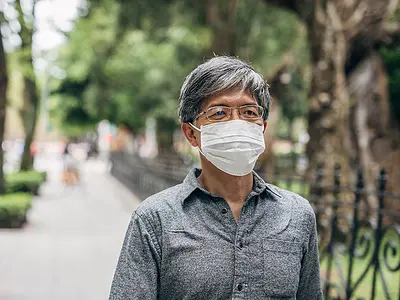 An Asian senior man wears a face mask and walks along a tree-lined sidewalk.