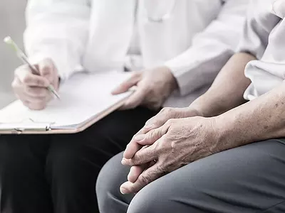 A medical professional in a white coat takes notes on a clipboard while talking to a senior patient.