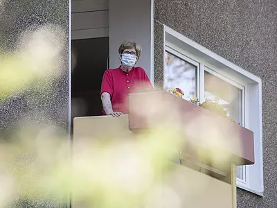 A senior woman wearing a mask looks out from a balcony.