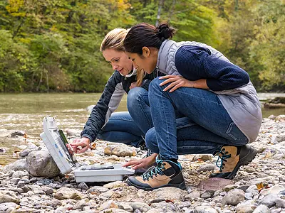 Two female scientists look at data on a tablet next to a creek.