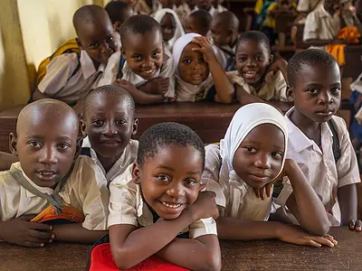 African schoolchildren crowd onto benches in a classroom and smile for the camera.