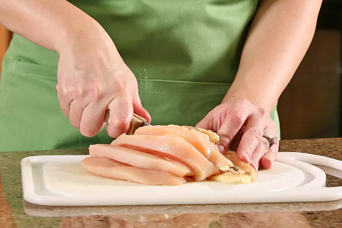A closeup of a woman slicing raw chicken breast on a cutting board.