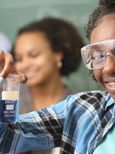A teenage African American girl holds a flask in a science classroom while classmates and a teacher work in the background.