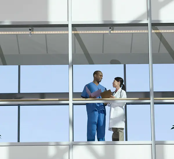 A male and female doctor talk in a glass-walled hospital walkway.