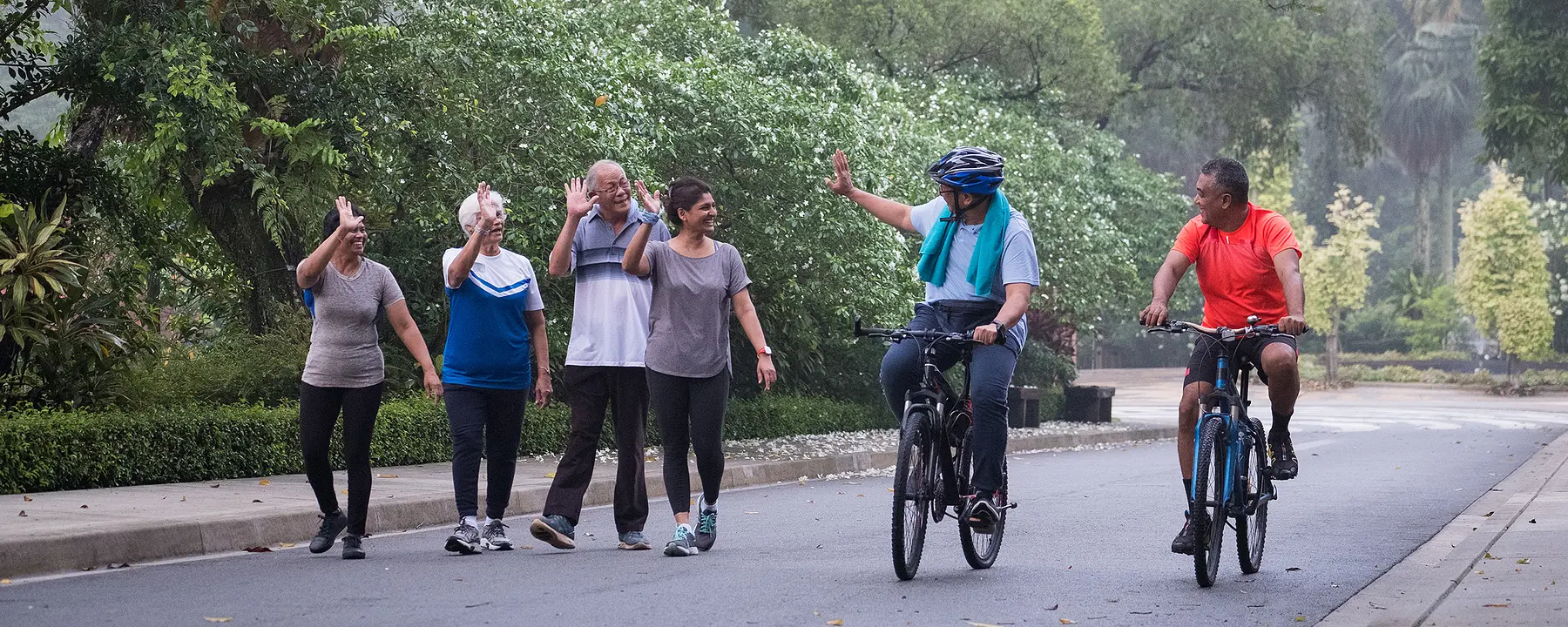 A group of adults walking in a city park wave at friends passing by on bicycles.