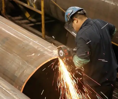 Worker using an angle grinder on a large metal pipe, surrounded by industrial equipment and pipes.