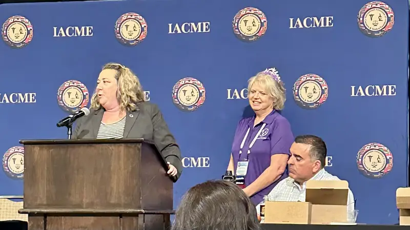 A woman speaks at a podium while two attendees listen and smile in a conference setting.